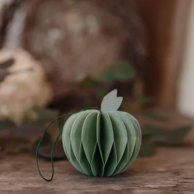 A green paper apple ornament with a string and a leaf on top, resting on a wooden surface with blurred foliage in the background.