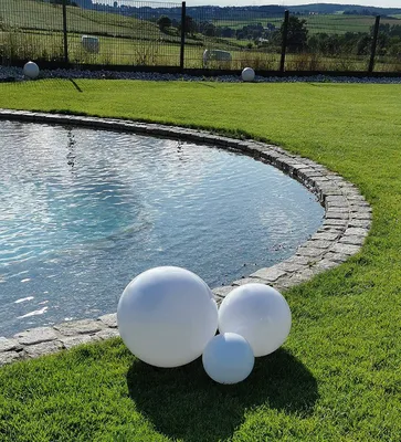 Three white spherical garden lights are placed on the grass next to a pond, with a fence and rolling hills in the background.