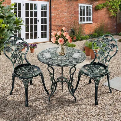 A bistro set with a round table and two chairs is placed on a gravel patio in front of a brick building with white double doors and a window.