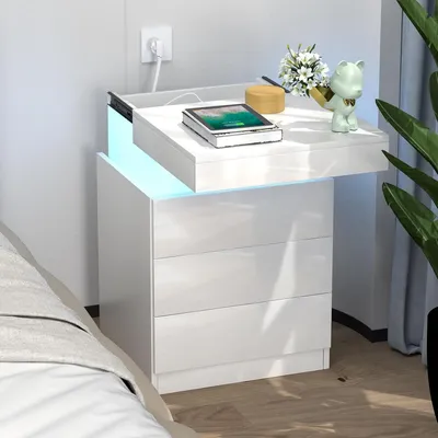 A white, high-gloss nightstand with three drawers, featuring a built-in LED light strip and a charging station on top. A tablet, a small decorative item, and a plant are placed on the nightstand.