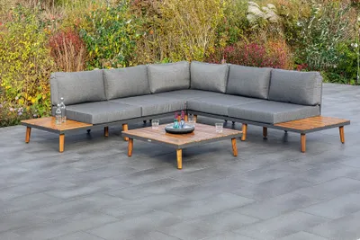 An outdoor seating set with a sectional sofa and coffee tables on a patio, with plants in the background.