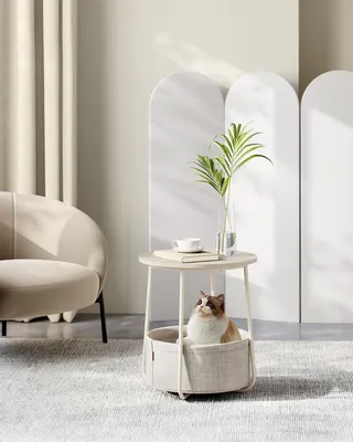 A cat is sitting in a round pet bed integrated into a side table, with a vase of greenery and a teacup on top. The scene is set in a modern, minimalist living room with soft lighting and neutral tones.