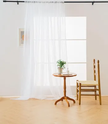 A sheer white curtain hangs in front of a window, with a small wooden table and chair to the side.
