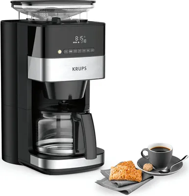 A black and silver Krups coffee maker with a glass carafe is shown on a white background, with a cup of coffee and two biscuits on a saucer to the right.