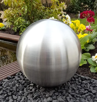 A metallic sphere sits on dark gravel in a garden setting, with plants and water visible in the background.