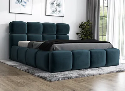 A dark blue upholstered bed with a tufted headboard and frame, placed on a grey rug in a room with artwork on the wall and a window with curtains in the background.