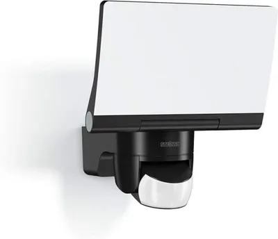 A black and white LED floodlight with a motion sensor is mounted on a white wall.