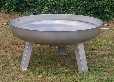 A stainless steel fire pit with legs and handles, placed on grass with a green hedge in the background.