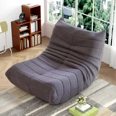 A gray floor chair with a textured fabric is placed on a rug in a room with a bookshelf and a window.