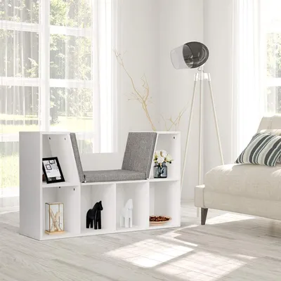 A white modular bookshelf with a built-in bench seat, decorated with various items, in a bright room with large windows and a floor lamp.