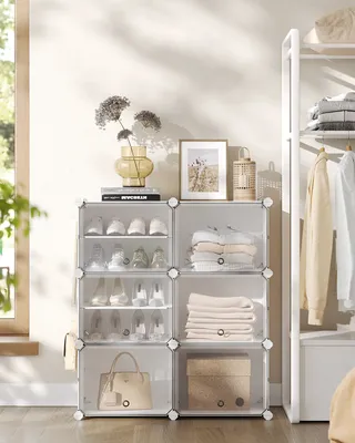 A white modular shelving unit filled with shoes, folded towels, and a handbag, positioned near a window and a tall white shelving rack with clothes.