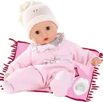A baby doll wearing a pink outfit and a white hat sits on a pink blanket with fringe.