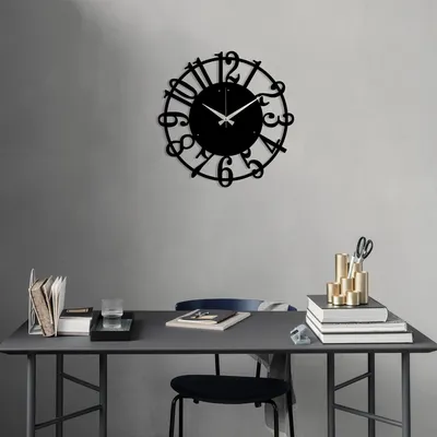 A black metal wall clock is mounted on a gray wall above a desk with various office supplies.