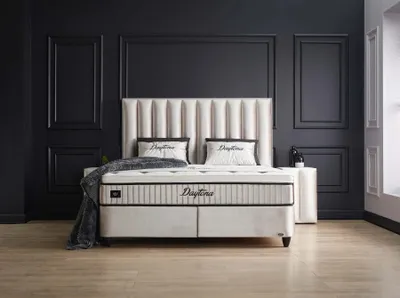 A bedroom scene featuring a large bed with a headboard, mattress, pillows, and a throw blanket. The room has dark gray walls with decorative molding and a light wood floor.