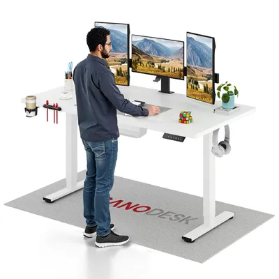 A man stands at a white standing desk with multiple monitors, a plant, and office supplies.