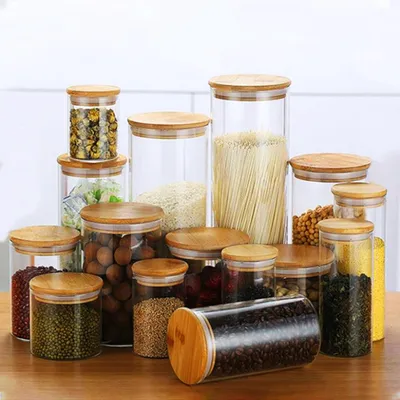 A collection of glass storage jars with bamboo lids filled with various dry goods like pasta, grains, beans, and coffee beans, arranged on a wooden surface.
