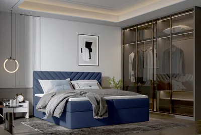 A bedroom scene featuring a dark blue boxspring bed with a headboard, pillows, and a blanket. A modern wardrobe with glass doors is visible on the right, and a nightstand with a lamp and alarm clock is on the left.
