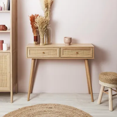 A console table with two drawers, decorated with dried flowers and a small pot, in a room with a pink wall and a jute rug.