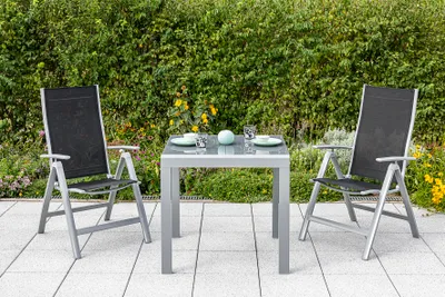 A garden table and two chairs set up on a patio with a green hedge in the background.