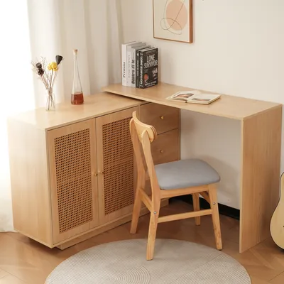 A light wood L-shaped desk with a cane-front cabinet and a gray upholstered chair in a room with sheer curtains.