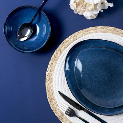 A table setting with blue plates, a bowl, and cutlery on a blue background, with a decorative flower in the corner.