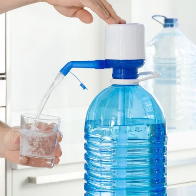 A hand is pressing down on a water dispenser pump attached to a large blue water bottle, dispensing water into a small glass. Another large water bottle is visible in the background.