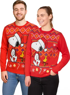 A man and a woman are posing for a product shot, both wearing matching red Christmas sweaters with Snoopy and Woodstock designs.