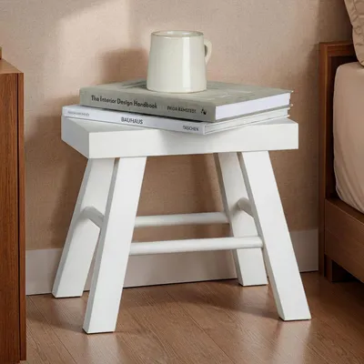 A white wooden stool with books and a mug on top, placed next to a bed.