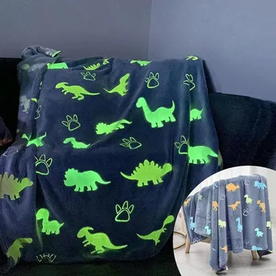 A glow-in-the-dark dinosaur blanket draped over a sofa, with a smaller inset image of the same blanket on a stool.