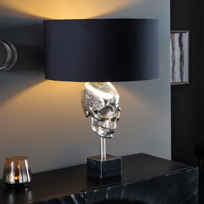 A table lamp with a skull base and a black lampshade sits on a dark cabinet.