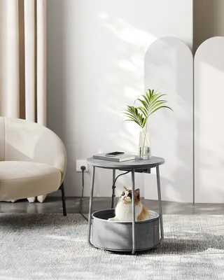 A cat is resting in a pet bed integrated into a side table, with a chair and curtains in the background.