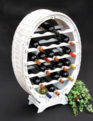 A white wooden wine rack shaped like a barrel, filled with wine bottles, placed on a black background with some greenery to the side.