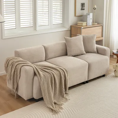 A beige sectional sofa with throw pillows and a blanket is placed in a living room setting, with a window and a wooden cabinet in the background.