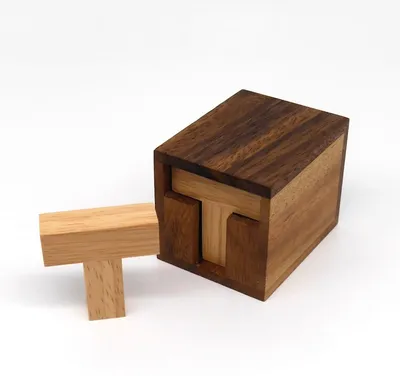 Holzbox mit dunklem Deckel, aus der ein helles T-förmiges Holzstück des „Six T“-Puzzles halb herausragt.