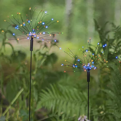 Two LED solar garden lights shaped like starbursts are placed in a garden setting with ferns and trees in the background.