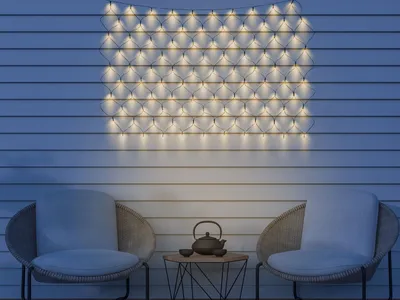 An outdoor patio scene at dusk, featuring two chairs, a small table with a teapot and cups, and a string of lights mounted on a wall.