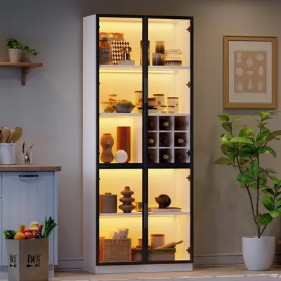 A tall, illuminated glass display cabinet filled with various items, placed in a room setting with a plant and a framed picture on the wall.