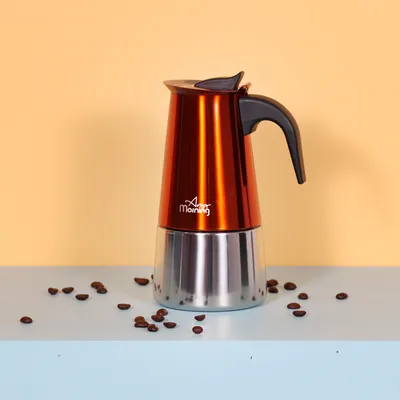 A copper-colored moka pot is placed on a light blue surface with coffee beans scattered around it.