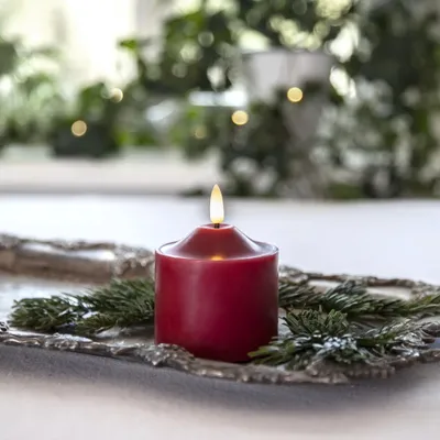 A red LED candle with a flame sits on a decorative tray with pine branches, with a blurred background of greenery and lights.