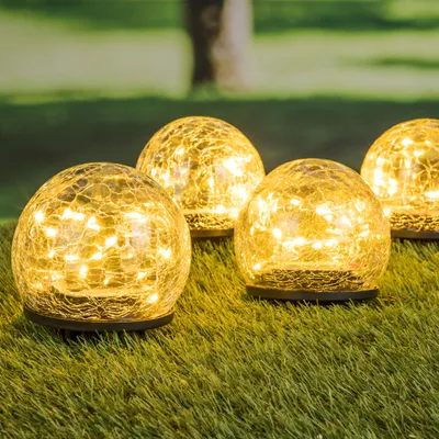 Four solar-powered crackle glass globe lights are arranged on a patch of green grass, with a blurred tree trunk and foliage in the background.