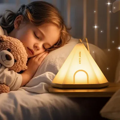 A young girl sleeping in bed with a teddy bear, next to a tent-shaped night light.