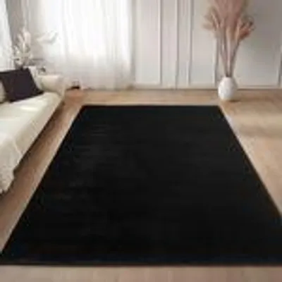 A black rug is placed on a wooden floor in a living room, next to a white sofa and in front of sheer white curtains.