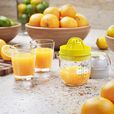 Two glasses of orange juice sit on a counter next to a manual juicer filled with orange juice. A bowl of oranges is in the background.