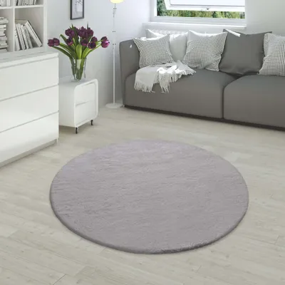 A round, gray rug is placed on a light wooden floor in a living room setting, with a gray sofa and white furniture visible in the background.