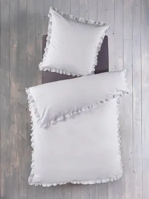 A top-down view of a white duvet cover and pillowcase with ruffled edges, laid out on a light gray wooden floor.