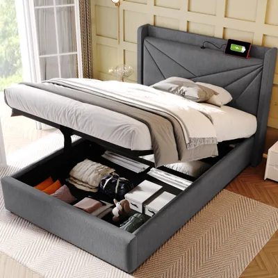 A grey upholstered bed with its storage compartment open, revealing folded items inside. The bed is in a room with a window and decorative wall paneling.