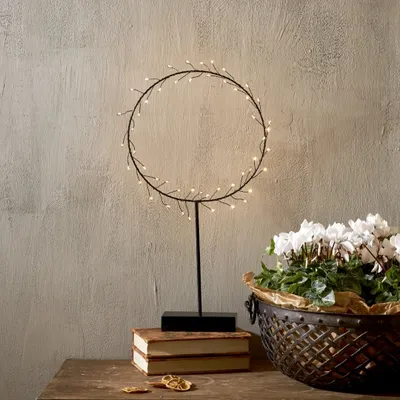 A decorative LED wreath light is displayed on a wooden table next to books and a basket of white flowers.
