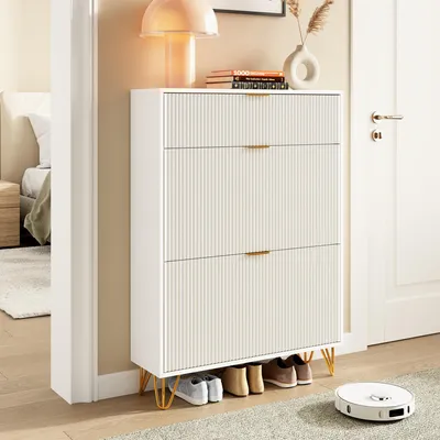 A white shoe cabinet with fluted drawers and gold legs, placed in a hallway, with shoes on the bottom shelf. A robotic vacuum cleaner is on the rug nearby.