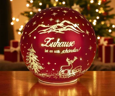 A red glass sphere with Christmas-themed decorations and text, placed on a wooden surface in front of a blurred Christmas tree and gifts.