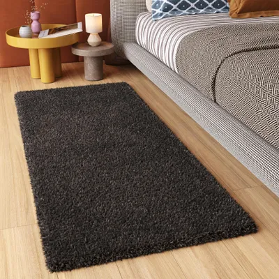 A dark gray rug is placed on a wooden floor next to a bed. A small side table with a lamp and decorative items is also visible.